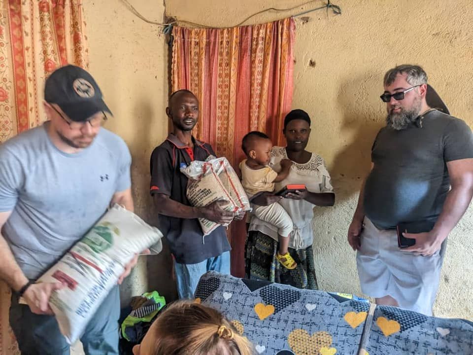 Men giving food to a family in a modest room. One man holds a bag, the other a child.