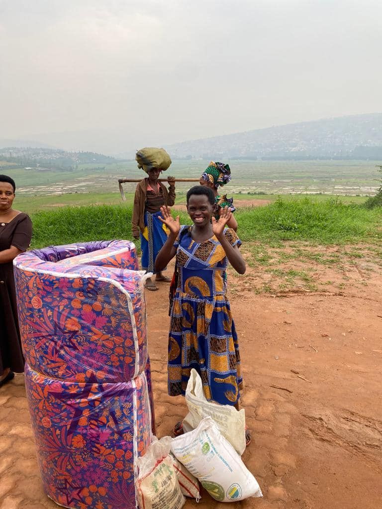 Woman in blue dress waves by rolled-up mattress, near bags and women carrying items on their heads in field.
