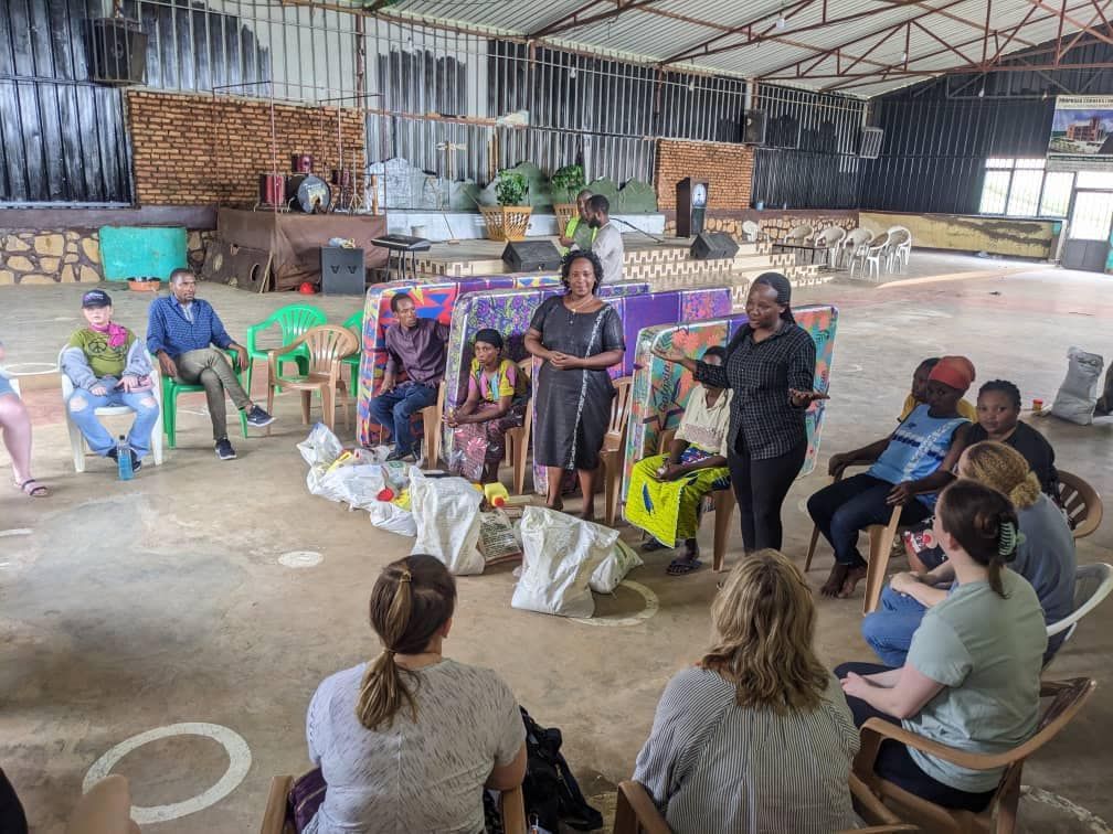 Group of people seated in a circle, listening to two women speaking in a large, open room.