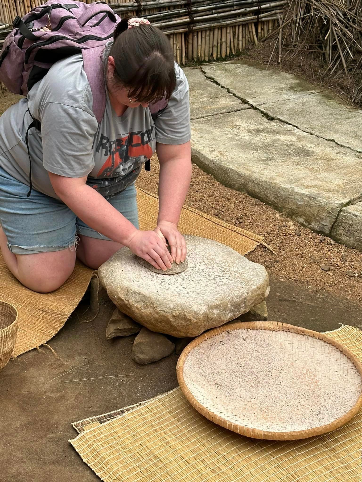 Woman kneeling, grinding grain on stone. Basket of grain nearby. Outdoor setting.