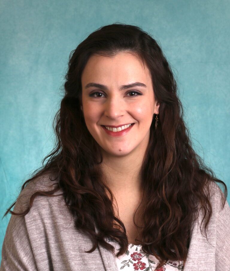 A woman with long brown hair is smiling in front of a blue background
