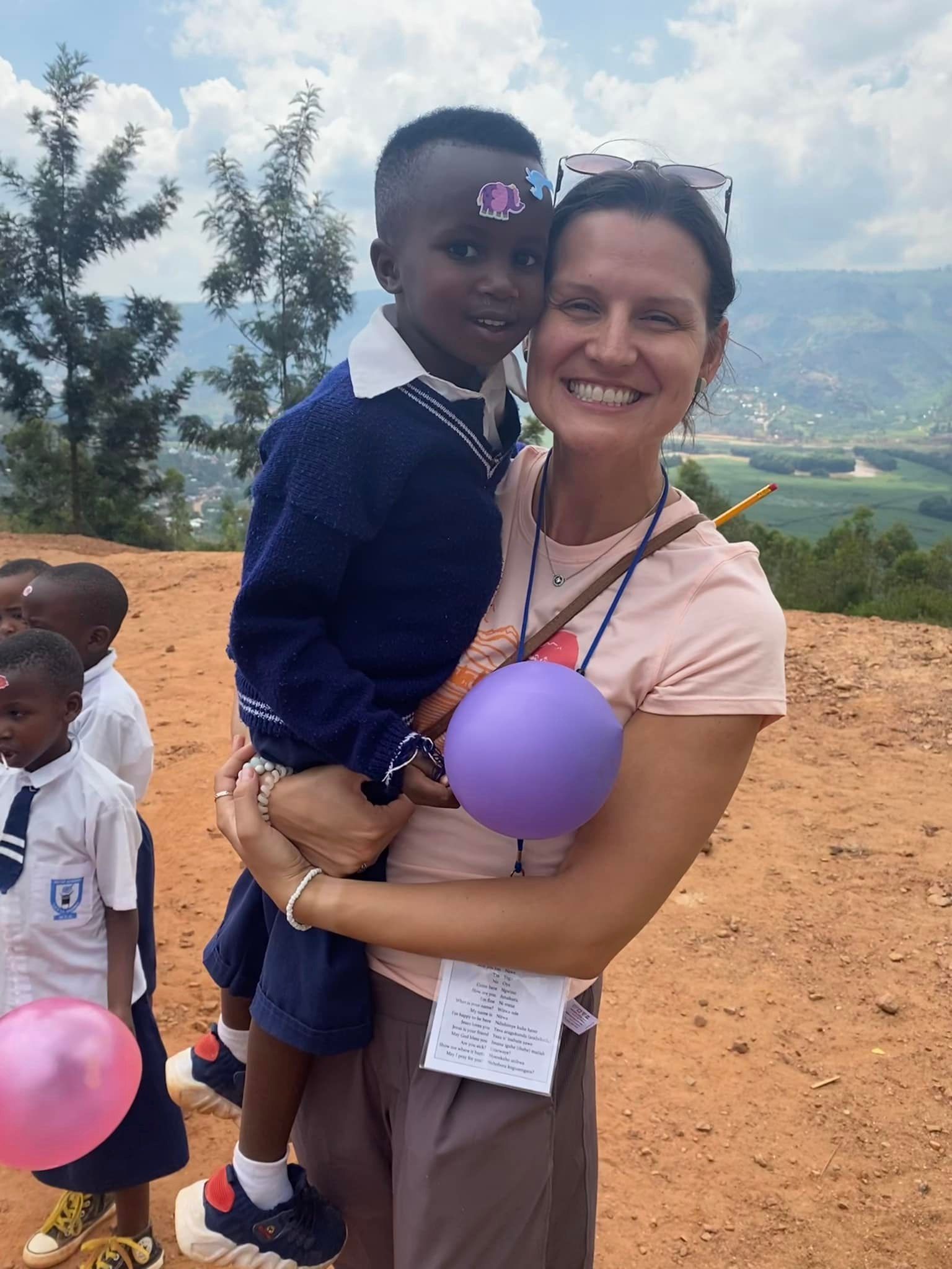 Woman holding a child, smiling, wearing a name tag, with other children in the background; outdoors, sunny day.