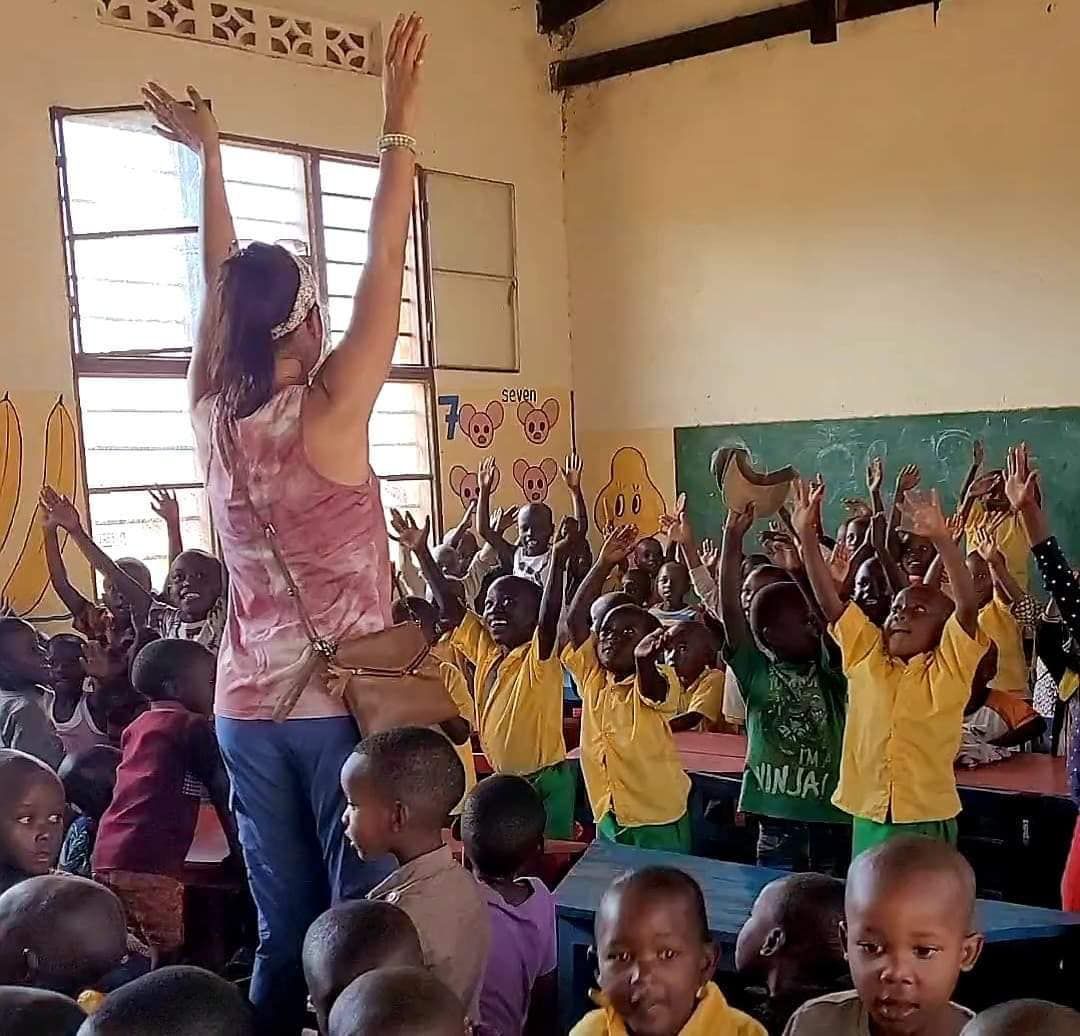 Woman in classroom with arms raised, surrounded by children raising hands.