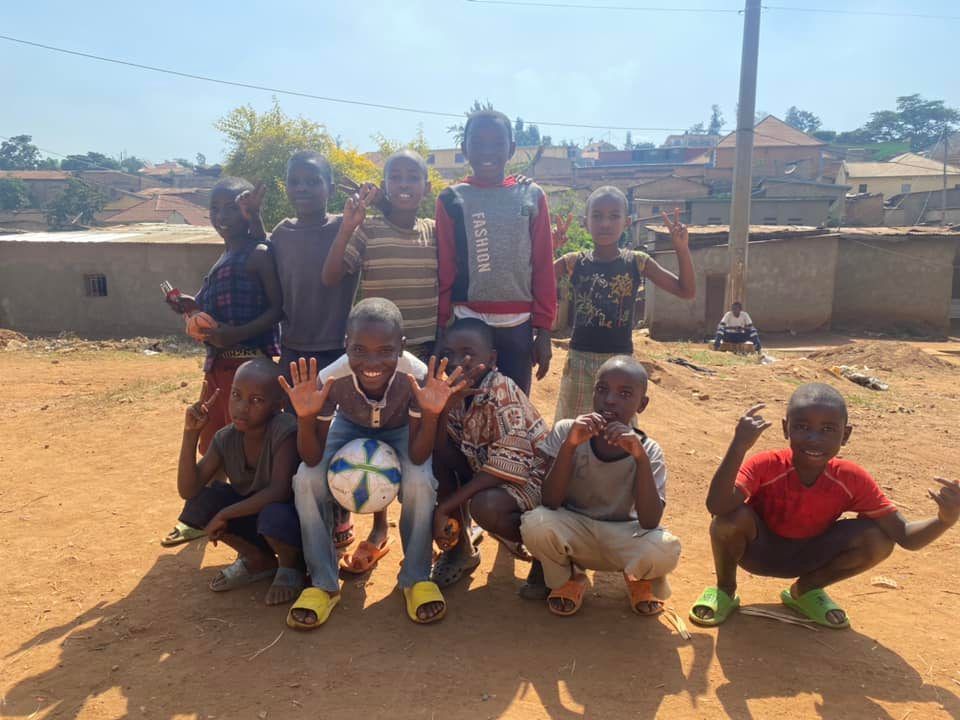 Group of smiling children posing outdoors with a soccer ball. They are waving and making hand gestures.