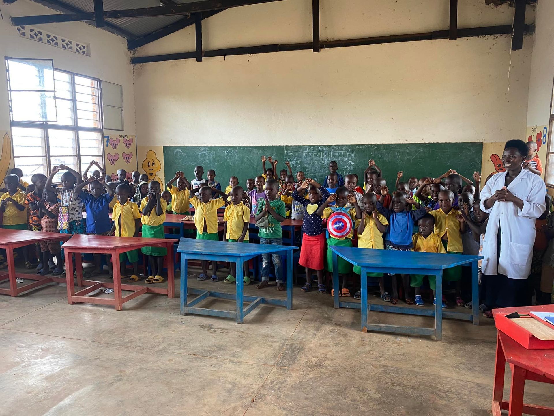 Children in colorful uniforms raising hands in a classroom; teacher stands near a chalkboard.