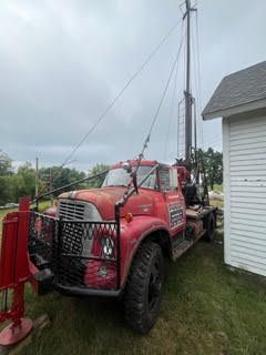 Red vintage well drilling truck parked near a white building, under a cloudy sky.