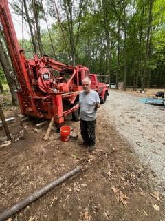 Man stands next to a red drilling rig in a wooded area. Bucket and metal pipe nearby.