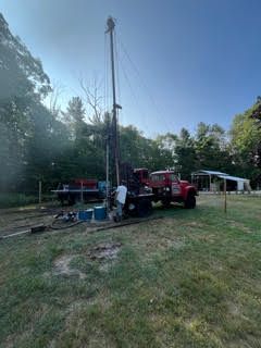 Red well drilling rig in a grassy field, near trees and a building, on a sunny day.