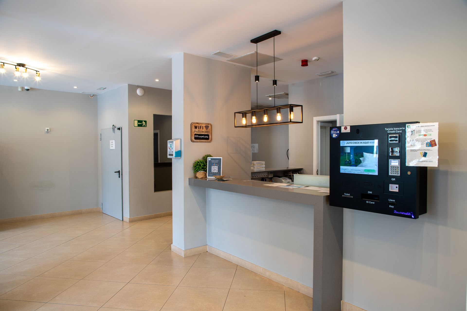 Vestíbulo de hotel con quiosco de registro y mostrador de recepción, luminoso, en tonos neutros y suelos de baldosas.