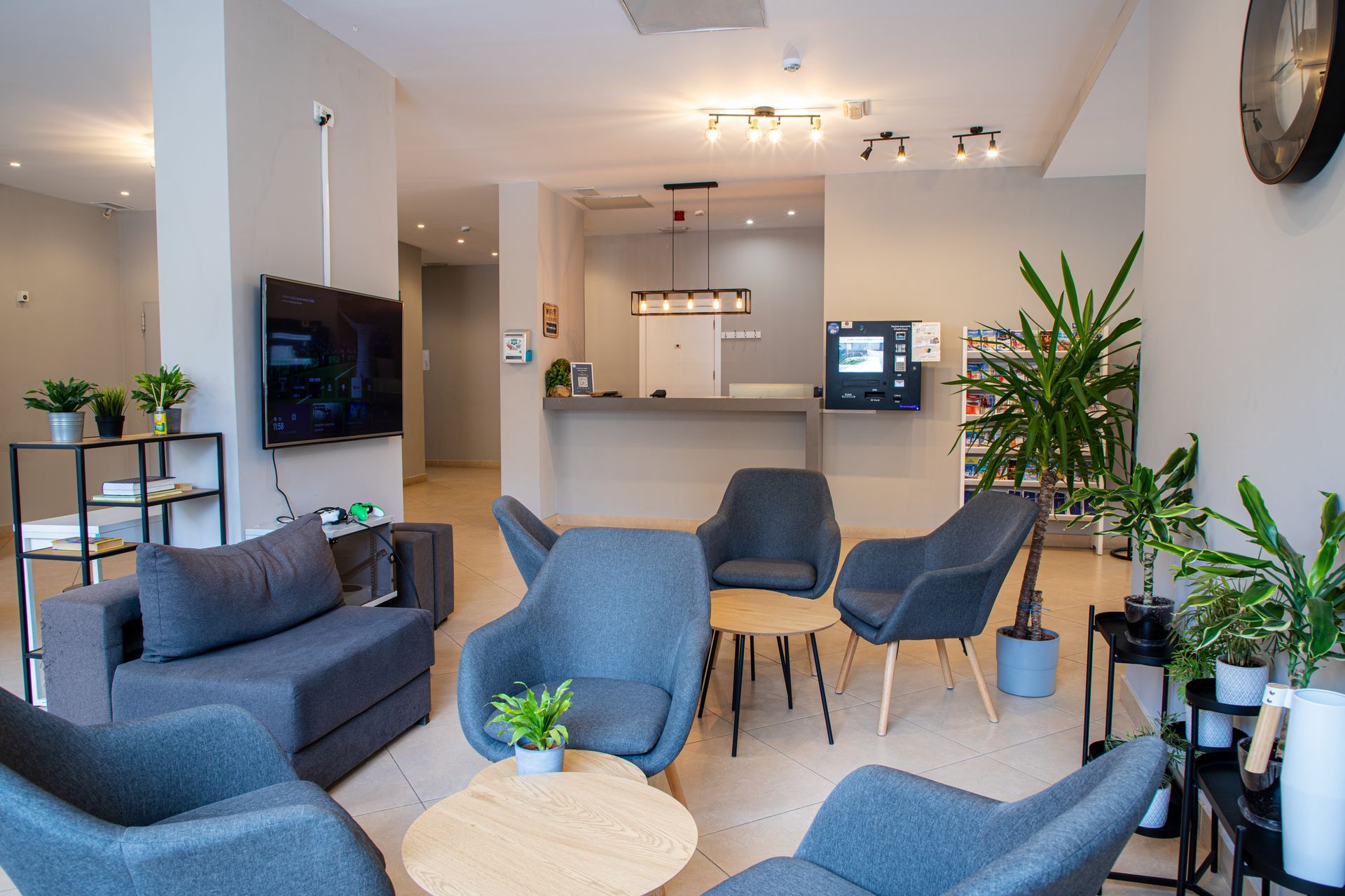 Zona de salón con sillones grises, mesas pequeñas, plantas, TV y un mostrador de recepción al fondo.