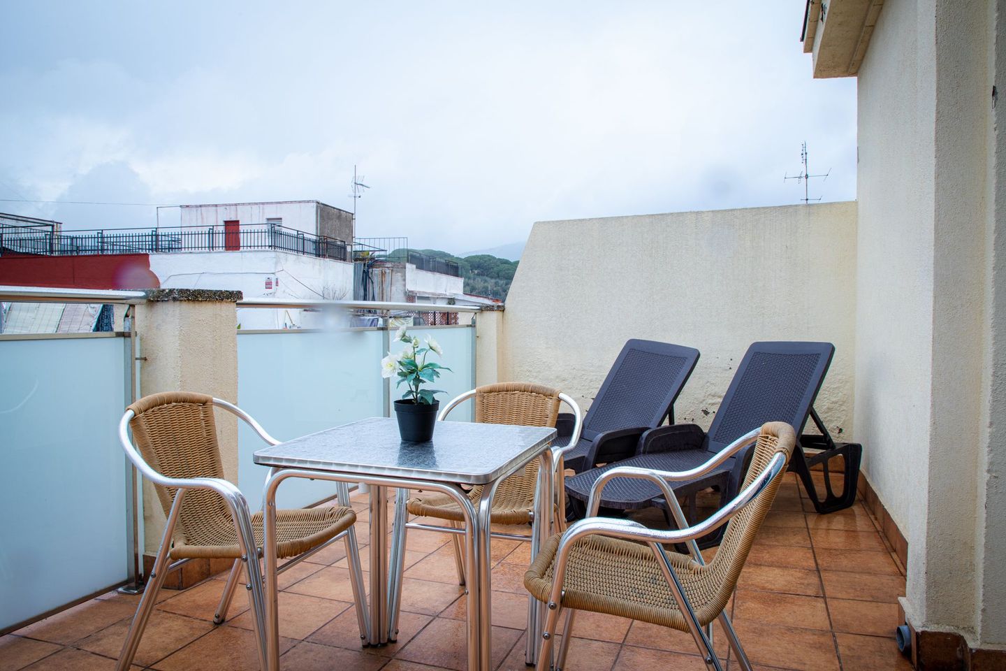 Patio en la azotea con mesa, sillas y tumbonas, con vistas a una ciudad en un día nublado.