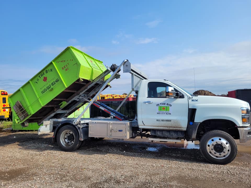 Green dumpster being lifted by a white roll-off truck on a sunny day.