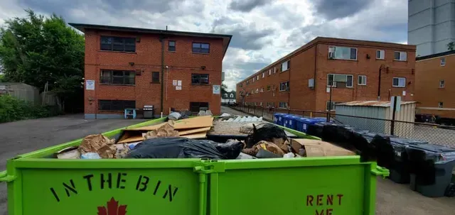 A green dumpster filled with trash in front of brick apartment buildings.