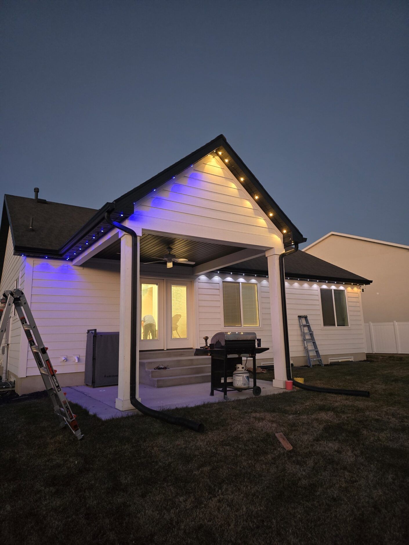 Small white house at dusk with blue and warm lights on the porch and roofline.