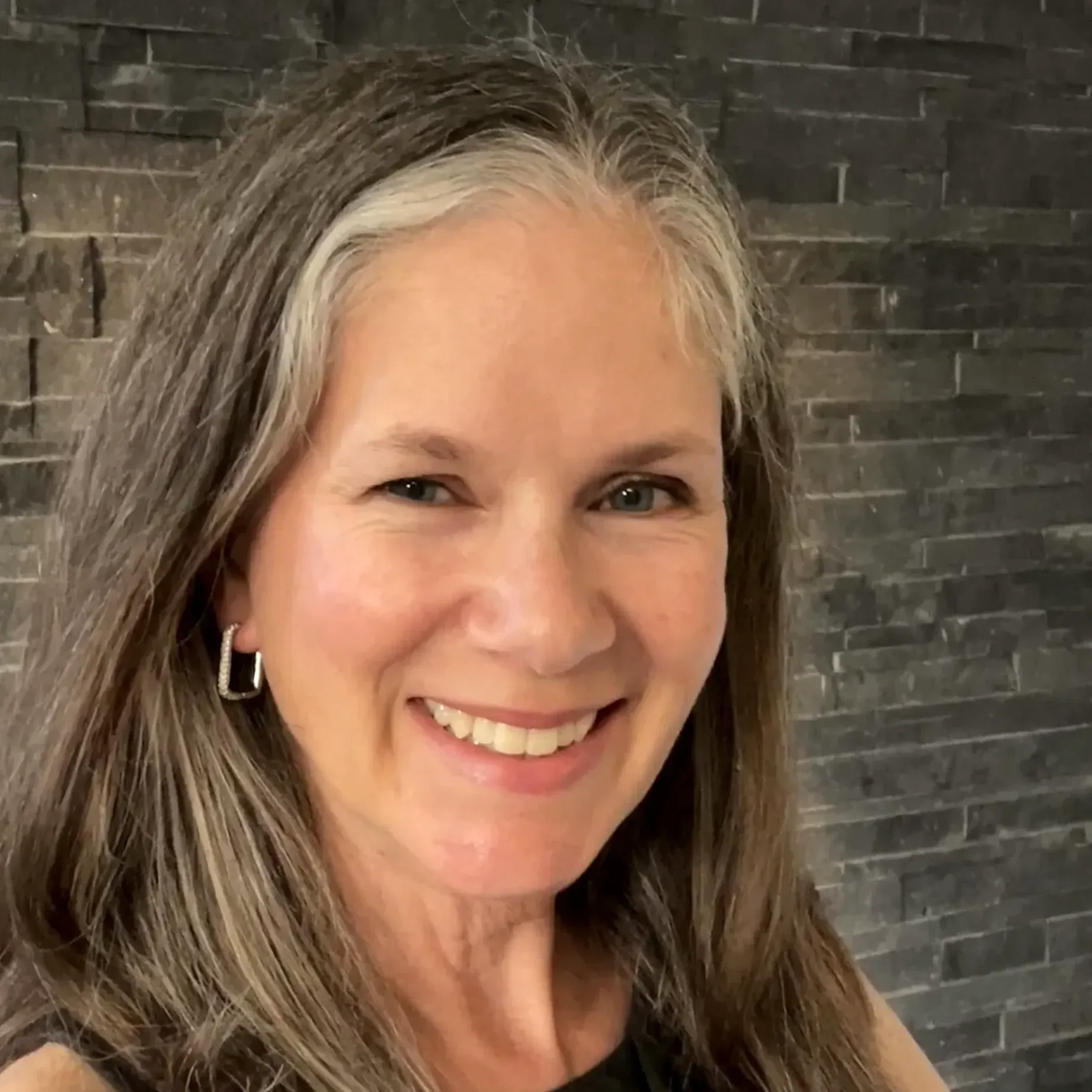 Smiling person with long hair featuring grey highlights, wearing rectangular earrings against a dark stone wall.