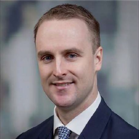 A person with short hair smiles in a professional headshot, wearing a dark blue suit, white shirt, and patterned tie.