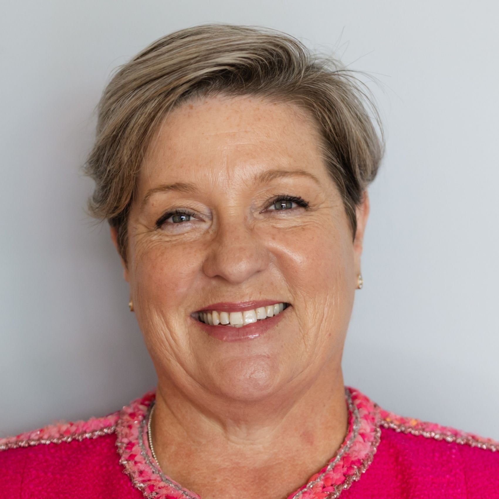 Headshot of a person with short, light brown hair smiling, wearing a bright pink textured top against a grey background.