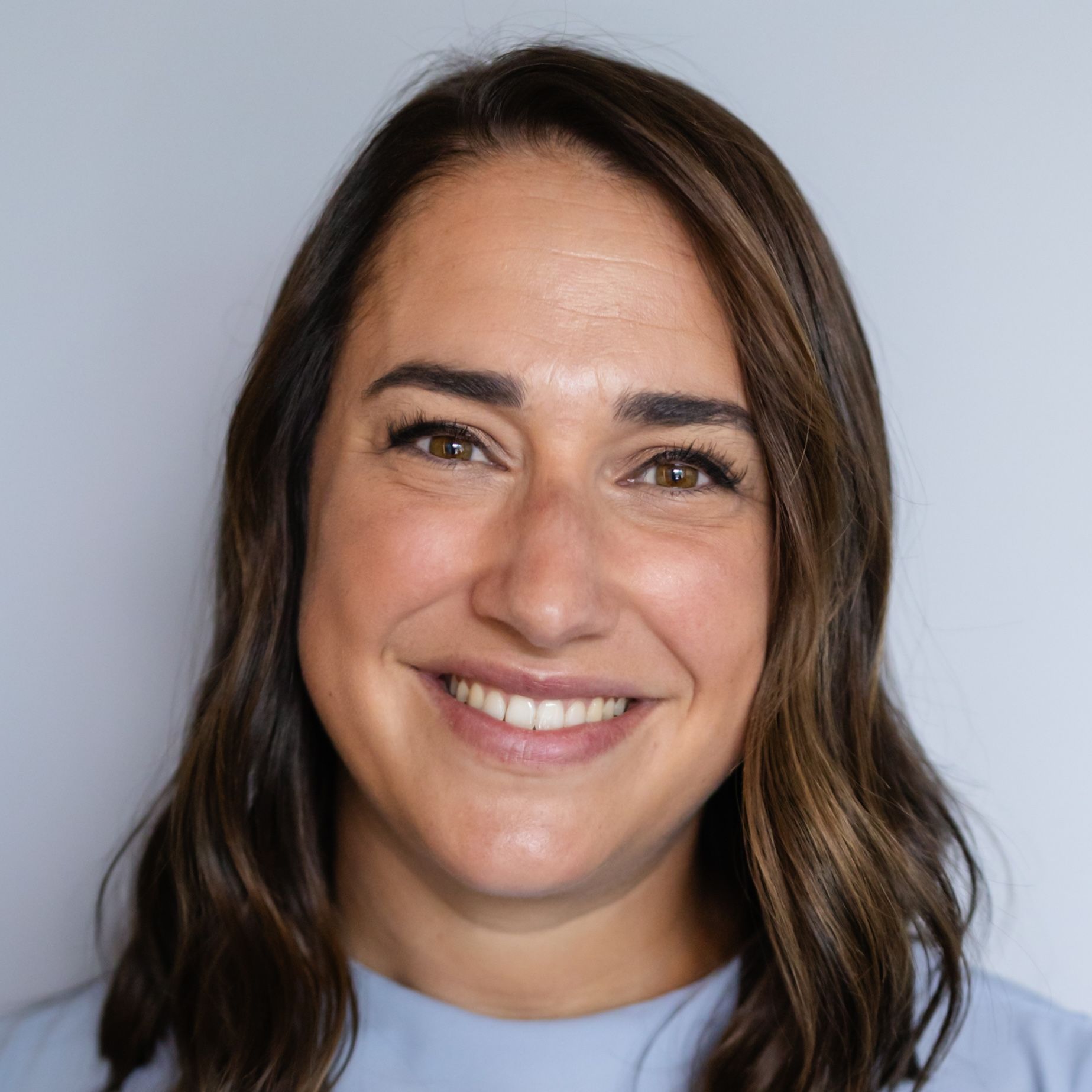 A smiling person with shoulder-length brown hair, wearing a light blue top against a plain light gray background.