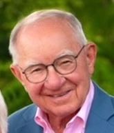 A smiling person wearing glasses, a pink collared shirt, and a blue blazer, set against a blurred green background.