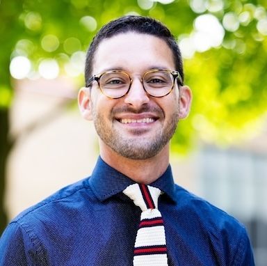 Smiling person with glasses wearing a navy blue button-down shirt and a white and red striped tie against a green background.