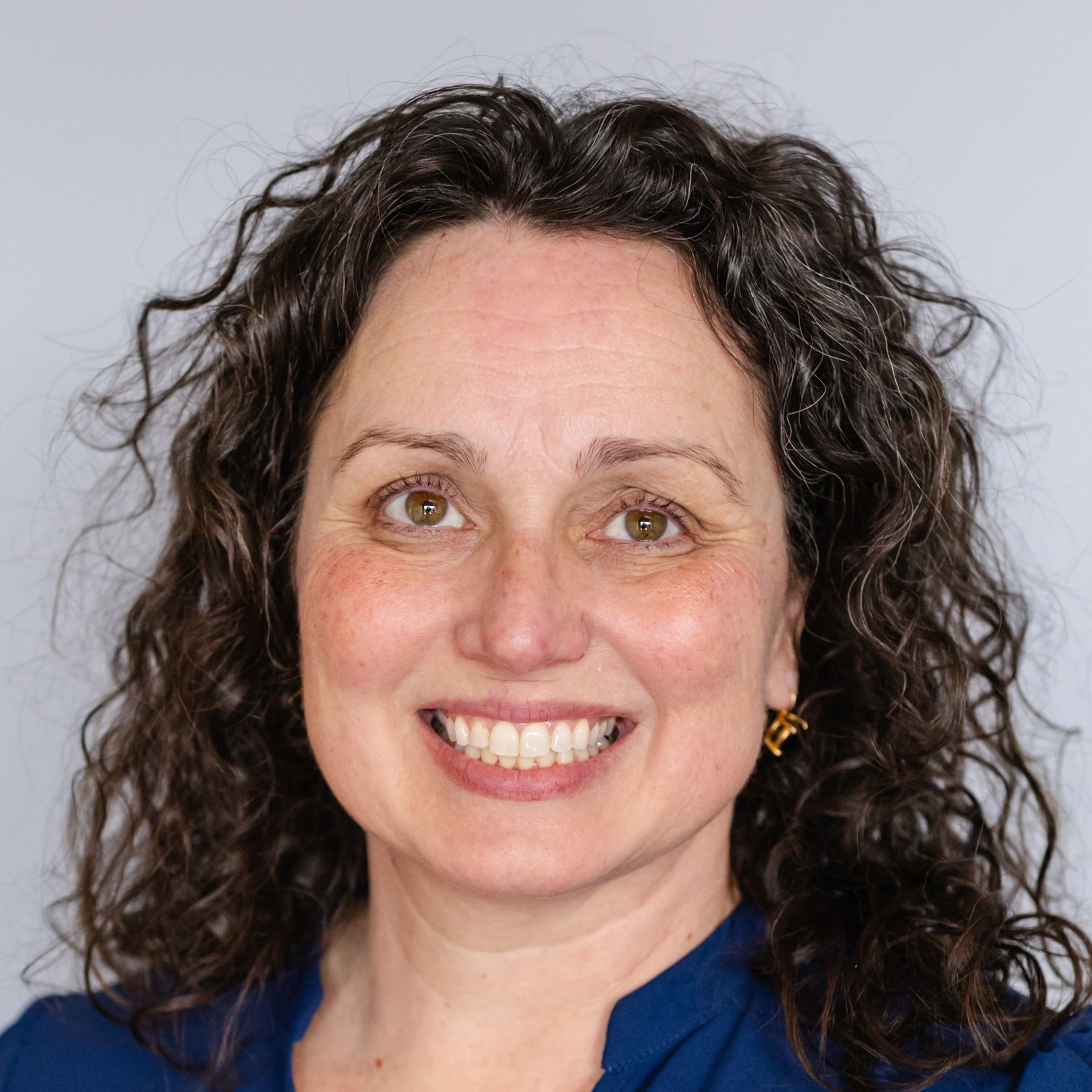 A person with curly brown hair and a bright smile wearing a blue top, set against a plain light gray background.