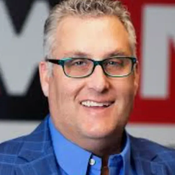 Smiling person wearing glasses and a blue plaid blazer against a background with red and white letters.