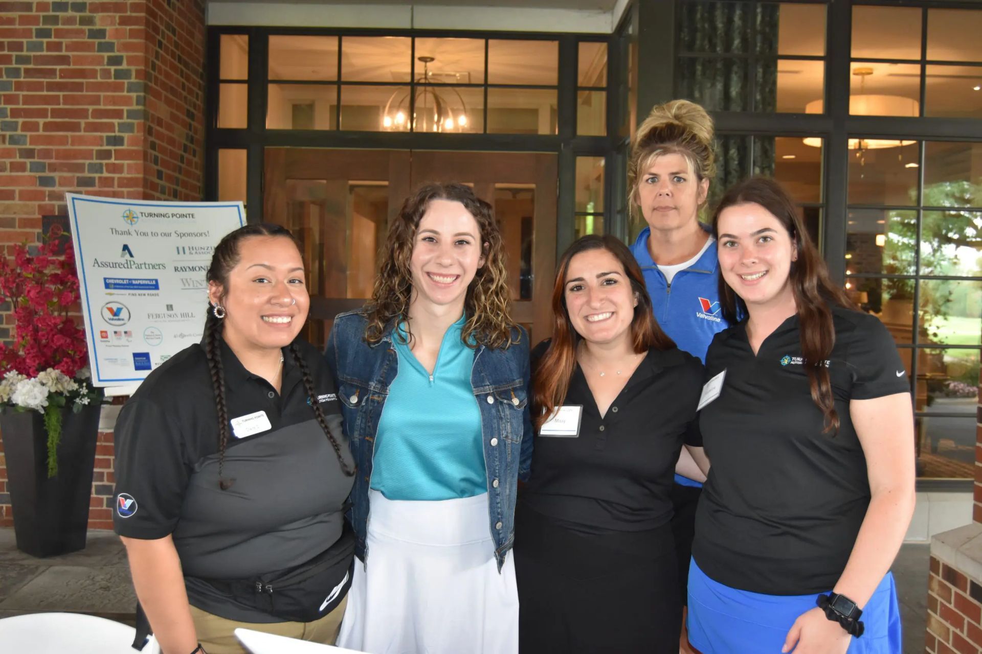 Five people stand together smiling outside a building entrance next to a sign for 