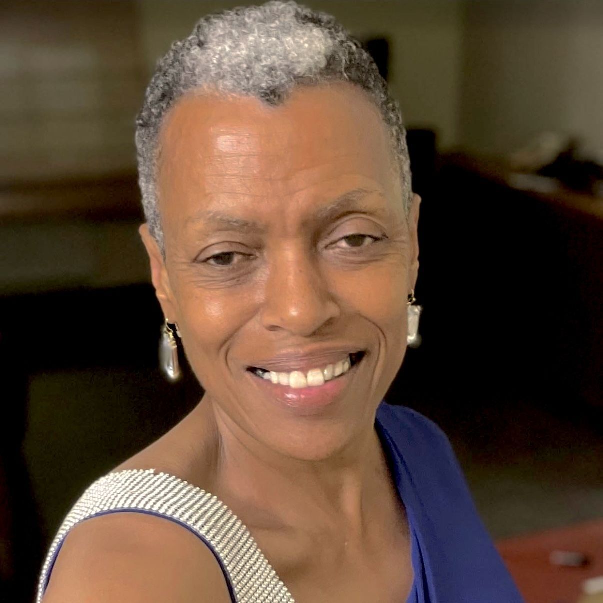 A person with short, grey hair and silver earrings smiles, wearing a blue top with a white beaded strap.