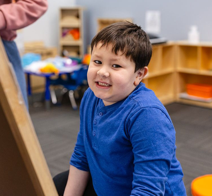 A smiling person in a blue long-sleeved shirt sitting in a classroom with wooden shelves in the background.