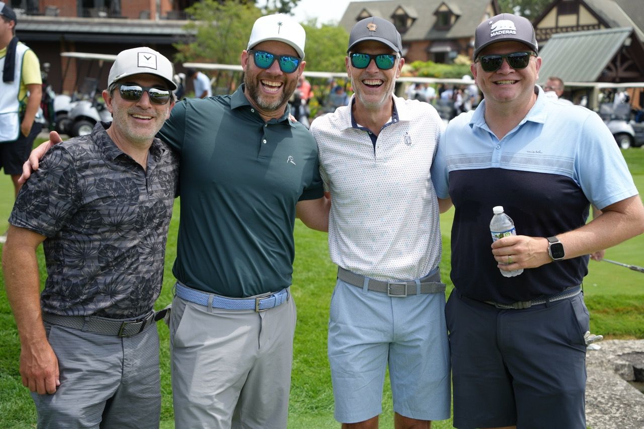Four men in golf attire and sunglasses smile together outdoors on a grassy golf course.