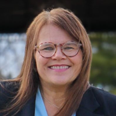 A person with shoulder-length brown hair, wearing glasses and a dark blue collared top, smiling against a light background.