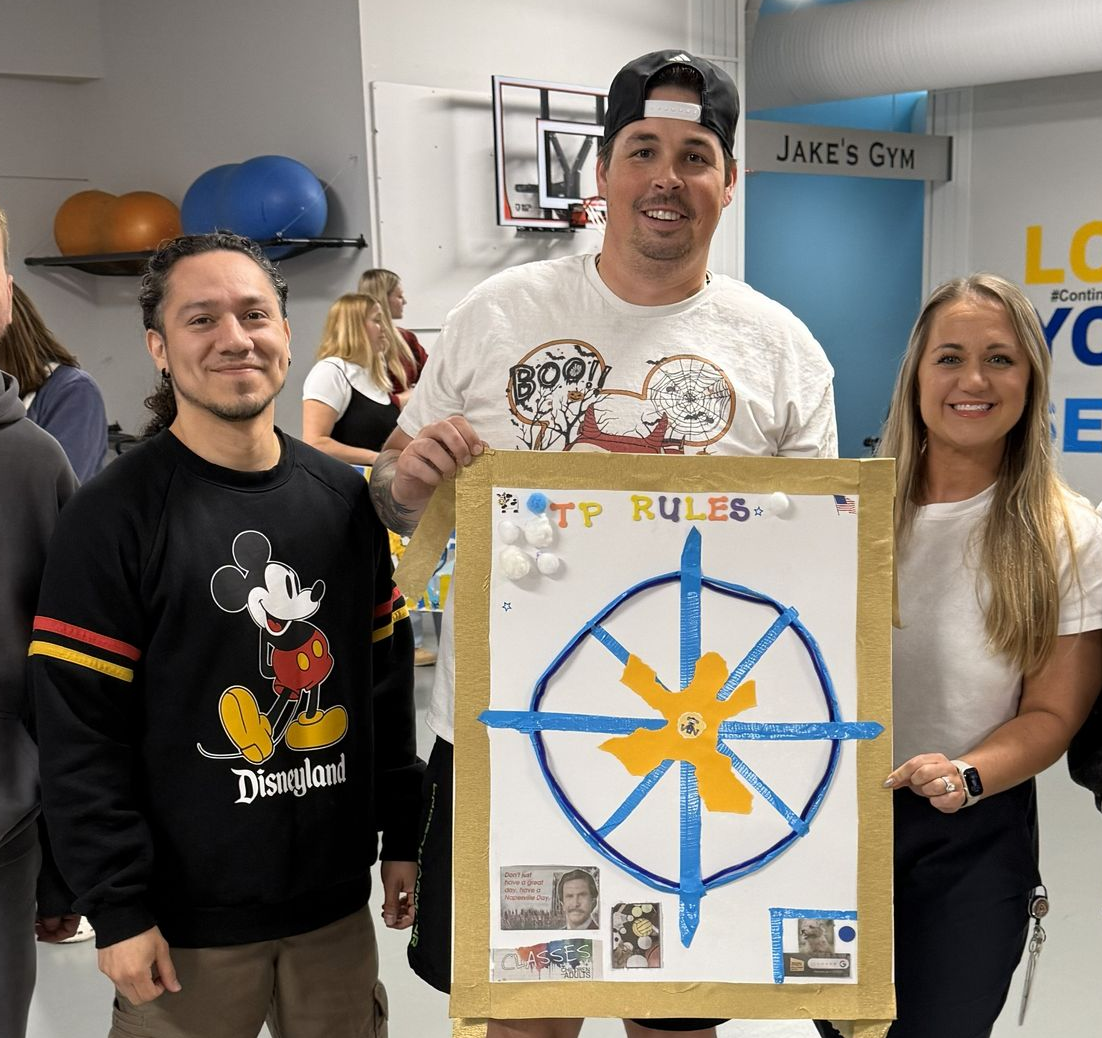 Three people pose in a gym holding a cardboard sign with a blue compass design, cotton balls, and printed photos.