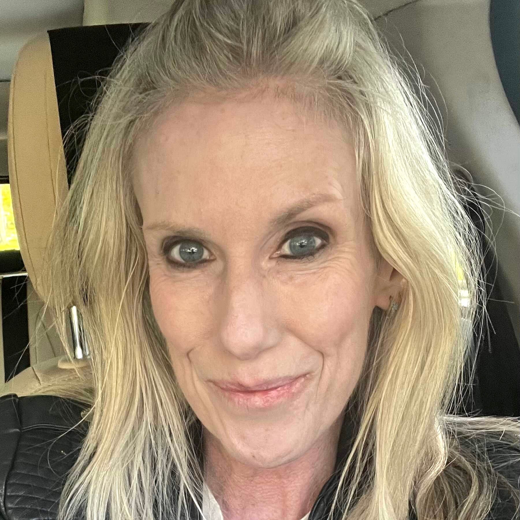 A selfie of a person with shoulder-length blonde hair and dark eyeliner, seated in a vehicle.