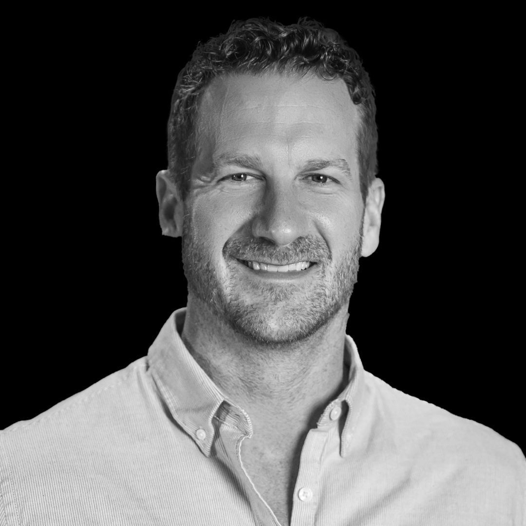 Headshot of a smiling person with short curly hair and a button-down shirt against a solid black background.