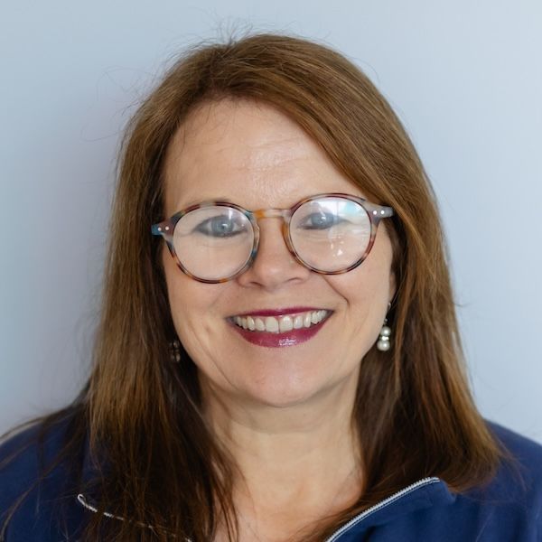 A person with shoulder-length brown hair, wearing glasses and a dark blue collared top, smiling against a light background.