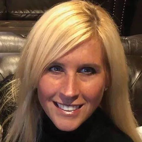 A person with long blonde hair and a friendly smile, centered in a close-up portrait against a dark, leather-like background.