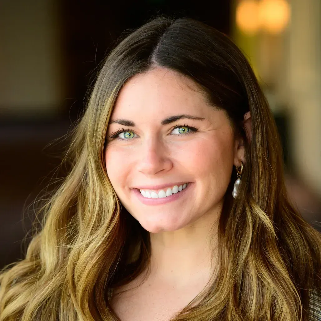 A headshot of a smiling person with long, highlighted brown hair and light green eyes, wearing simple drop earrings.