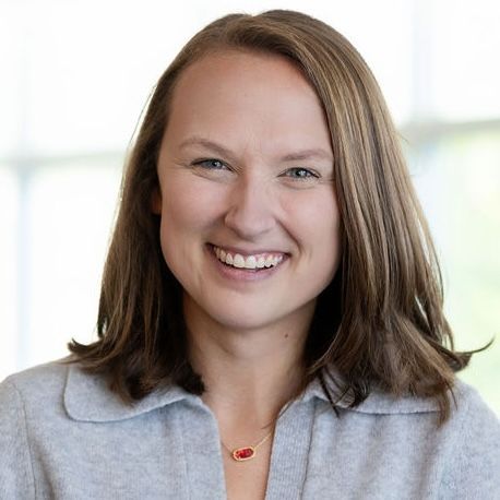 Smiling person with shoulder-length brown hair, wearing a gray collared shirt and a delicate gold necklace with a red charm.