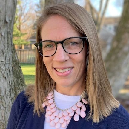 A smiling person with shoulder-length blonde hair and glasses, wearing a white shirt, blue cardigan, and pink necklace.