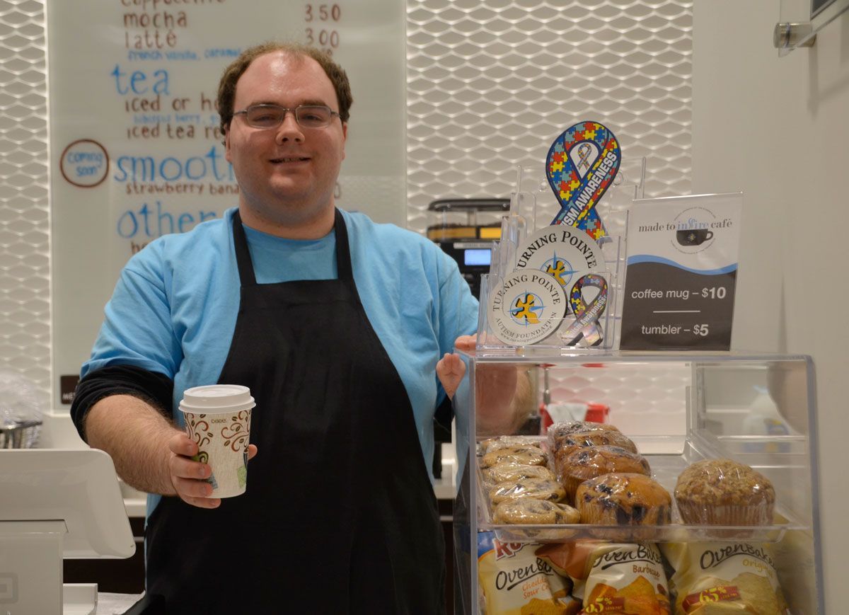 A barista in a blue shirt and black apron holds out a paper coffee cup next to a display of pastries in a cafe.