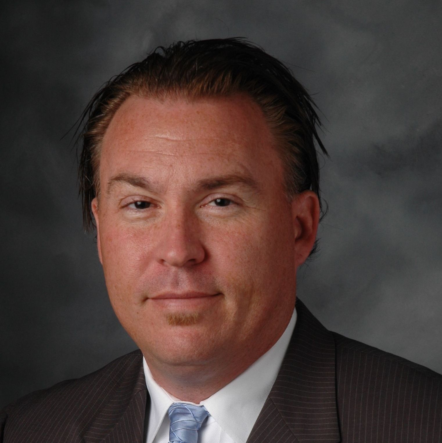 Professional headshot of a person in a brown suit, white shirt, and blue tie against a dark grey mottled background.