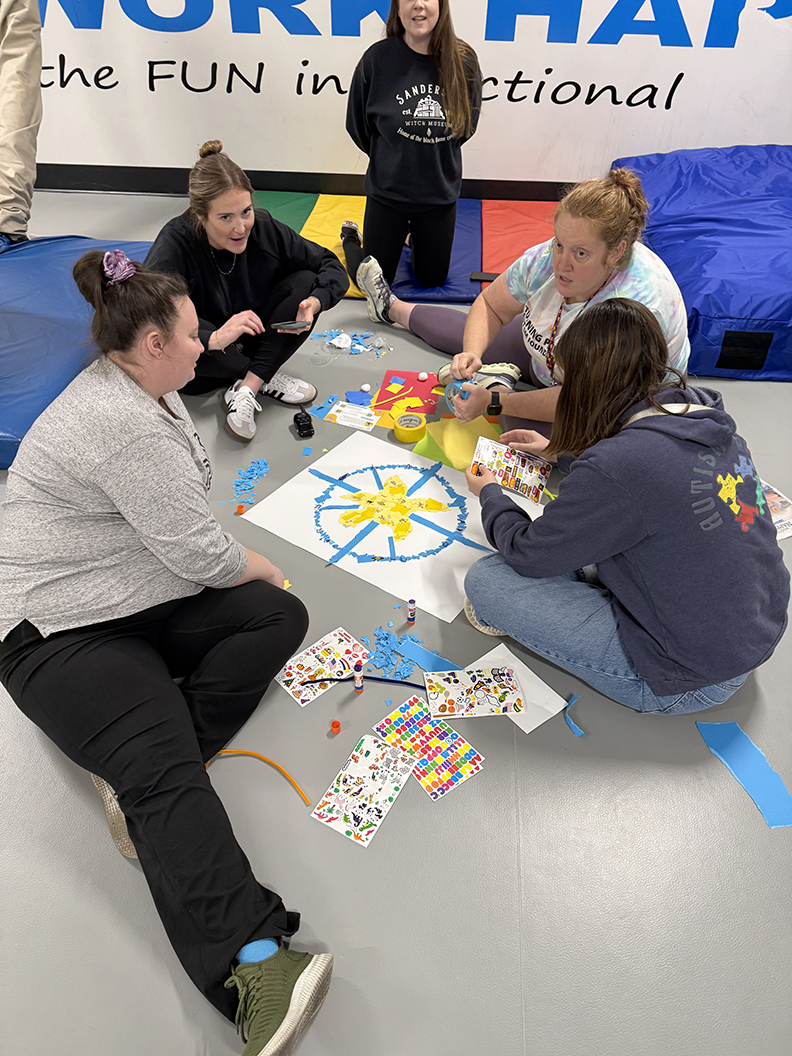 Four people sit on a gym floor collaborating on an art project featuring a central star design on a white poster.