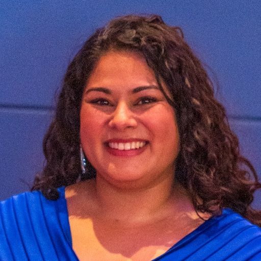 A smiling person with long dark wavy hair wearing a royal blue top against a blue background.