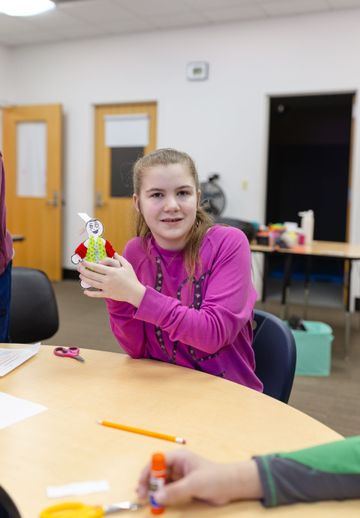 A person in a pink shirt sits at a desk holding a small, handmade craft figure with a white head and colorful body.