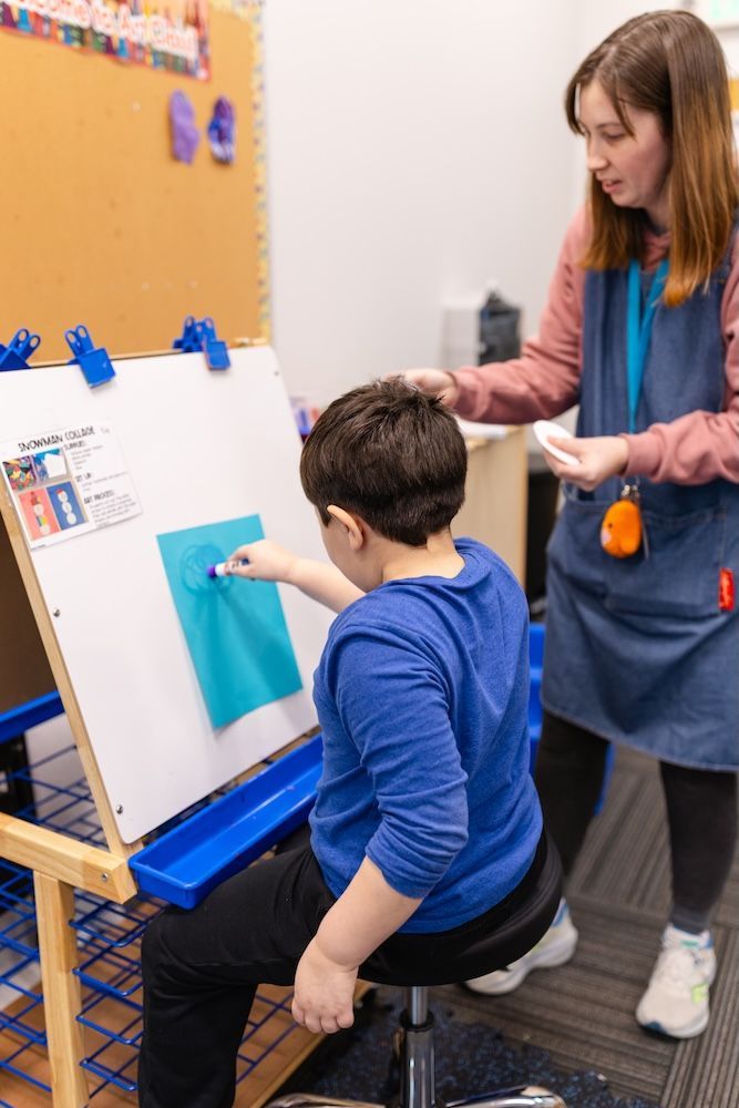 A teacher guides a child as they paint a blue circle on a blue sheet of paper attached to an easel in a classroom.