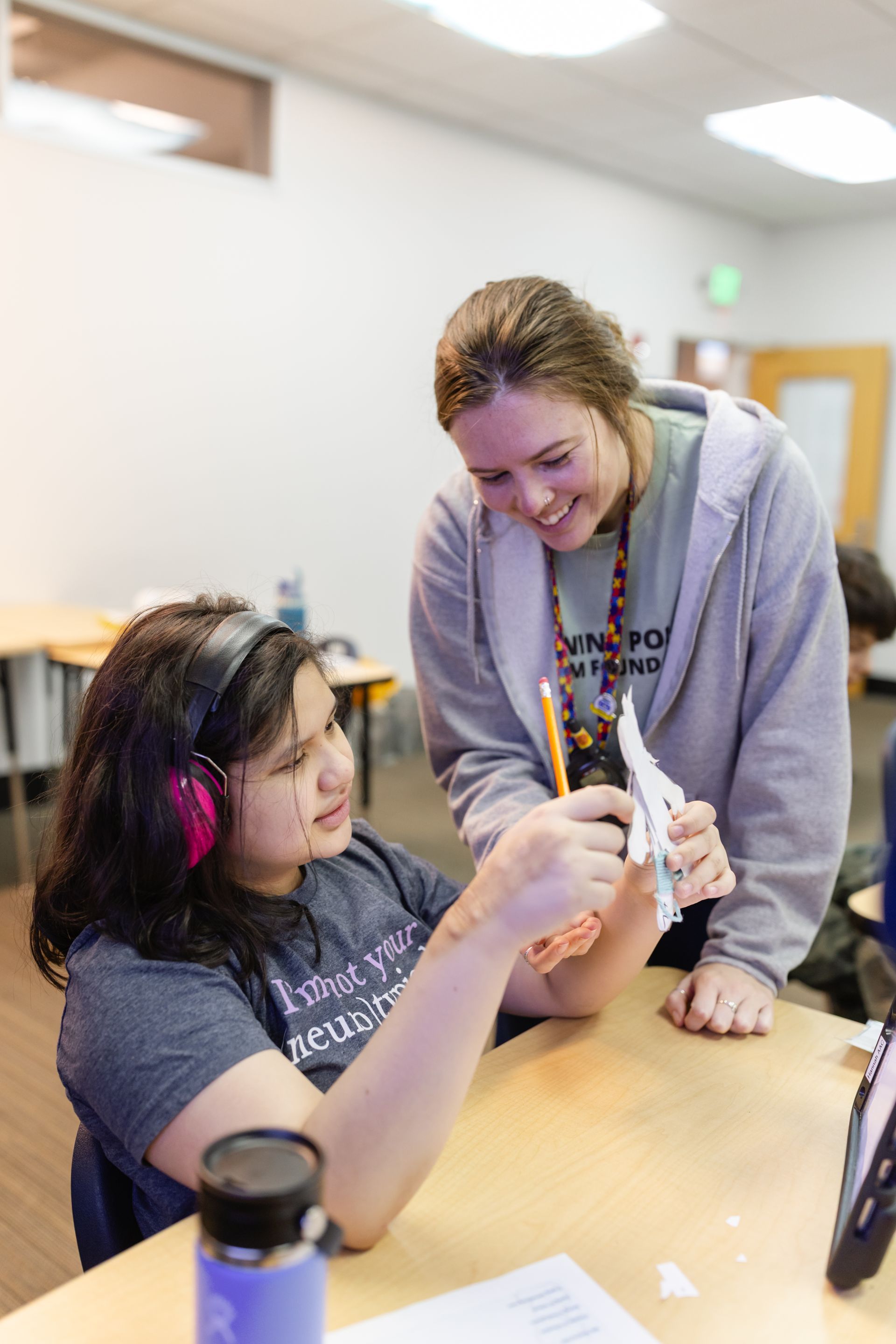 A student with headphones and an instructor look at a paper model together at a desk in a classroom.