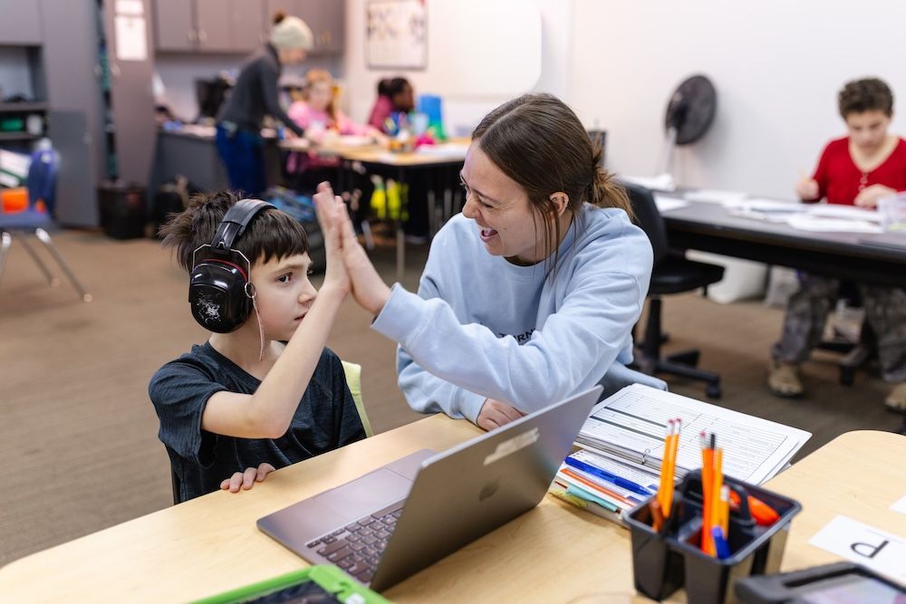A student wearing headphones high-fives a teacher across a desk with a laptop in a classroom.