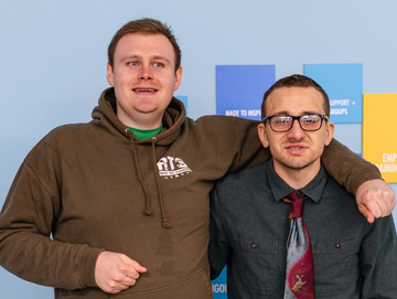 Two people pose side-by-side against a blue wall with colored signage; one wears a brown hoodie, the other a tie and shirt.