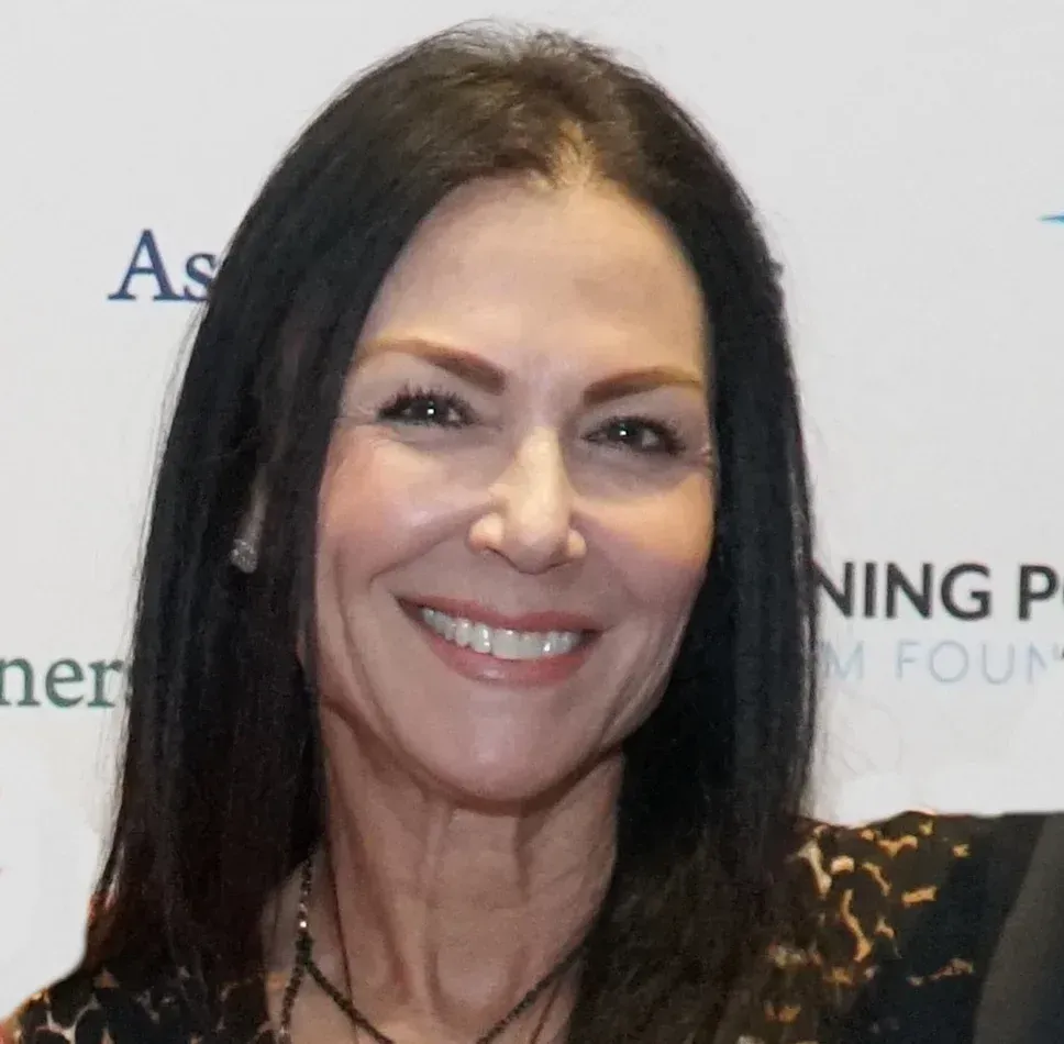 A woman with long dark hair smiles warmly in front of a partially visible branded event backdrop.
