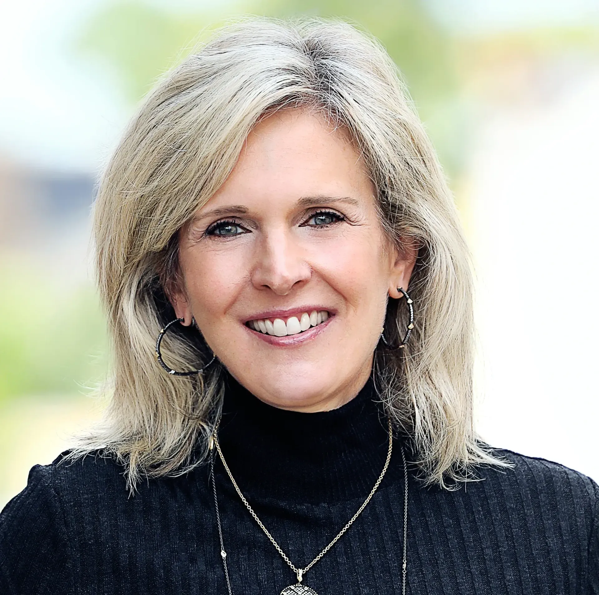 A smiling woman with blonde hair, wearing a black turtleneck and necklace, against a soft-focus background.